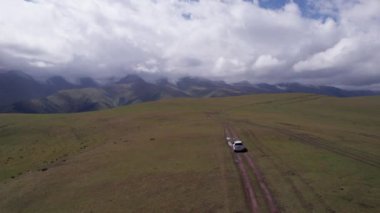 Beyaz bir SUV dağlarda tarlalardan geçiyor. Devasa beyaz bulutlardan, karlı dağ zirvelerinden ve yeşil tarlalardan oluşan bir dronun hava görüntüsü. Su birikintileri olan kirli bir off-road. Tekerleklerden damlıyor