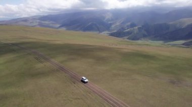 Beyaz bir SUV dağlarda tarlalardan geçiyor. Devasa beyaz bulutlardan, karlı dağ zirvelerinden ve yeşil tarlalardan oluşan bir dronun hava görüntüsü. Su birikintileri olan kirli bir off-road. Tekerleklerden damlıyor