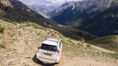 Beyaz bir SUV nehir boyunca uzanan bir vadi boyunca bir dağ yolunun üstesinden gelir. Uçurumlu yeşil alanlar. Dik uçurumlar ve yoldaki büyük kayalar. Keskin bir iniş ve tepe manzarası. Noel ağaçları