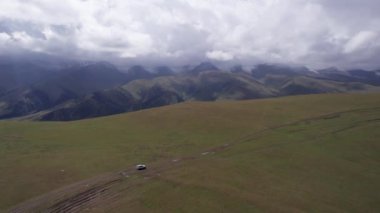 Beyaz bir SUV dağlarda tarlalardan geçiyor. Devasa beyaz bulutlardan, karlı dağ zirvelerinden ve yeşil tarlalardan oluşan bir dronun hava görüntüsü. Su birikintileri olan kirli bir off-road. Tekerleklerden damlıyor
