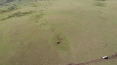 Beyaz bir SUV dağ yeşili tarlalardan geçiyor. Uzakta karlı zirveleri olan yüksek tepeler var. Kirli yol. Mavi gökyüzü. Bir köpek koşar ve çobanlık yapar. Dağlık arazi, bazen orman.