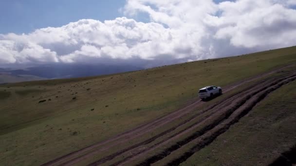 一辆白色的越野车在山上穿过田野 空中俯瞰着巨大的白云 雪峰和绿地 肮脏的越野车和水坑 车轮上的水花 — 图库视频影像