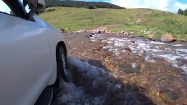 Beyaz bir SUV nehirden ve çamurdan geçiyor. Tekerleklerin manzarası. Beyaz boya etrafındaki dünyayı yansıtıyor. Nehirden sular, kayalar, dağlı ve ormanlı yeşil çimenler. Mavi gökyüzü
