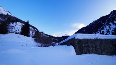 An old stone prison or hut in the mountains. Snowy mountains, forest and clouds against a blue sky. Tall dry bushes peek out from under the snow. Birch and spruce trees are visible in the distance.