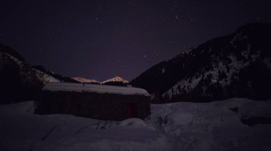Gece yıldızlı gökyüzü ve karlı dağlarda bir kulübe. Takımyıldızları görülebilir, yıldızlar parlak. Uzakta karlı tepelerin ve ormanların siluetleri. Eve giden bir yol var.