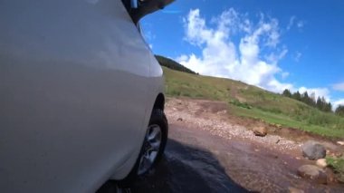 Beyaz bir SUV nehirden ve çamurdan geçiyor. Tekerleklerin manzarası. Beyaz boya etrafındaki dünyayı yansıtıyor. Nehirden sular, kayalar, dağlı ve ormanlı yeşil çimenler. Mavi gökyüzü