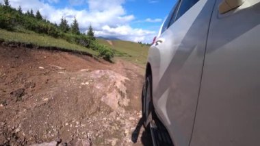 Beyaz bir SUV nehirden ve çamurdan geçiyor. Tekerleklerin manzarası. Beyaz boya etrafındaki dünyayı yansıtıyor. Nehirden sular, kayalar, dağlı ve ormanlı yeşil çimenler. Mavi gökyüzü