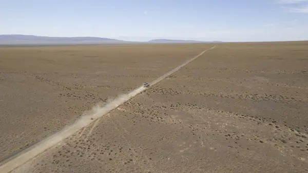 stock image A white SUV is driving fast on a dusty road in the steppe. Clouds of dust fly from under the wheels and from the roof of the car. The jeep easily passes a dirt road. Blue sky and white clouds.