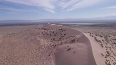 Bozkırın ortasında büyük bir kum tepeciği. Kocaman bir kum yığınının drone görüntüsü. Turistler yürüyor, manzaranın tadını çıkarıyorlar. Uzakta bir nehir akar ve çimenler büyür. Beyaz bulutlu gökyüzü