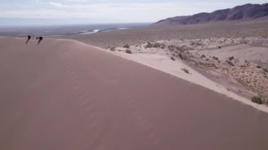 Bozkırın ortasında büyük bir kum tepeciği. Kocaman bir kum yığınının drone görüntüsü. Turistler yürüyor, manzaranın tadını çıkarıyorlar. Uzakta bir nehir akar ve çimenler büyür. Beyaz bulutlu gökyüzü