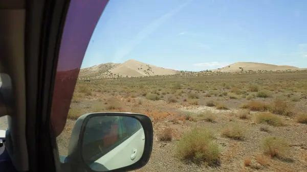 View from the car interior in the side mirror. A white car is moving along a dirt road in the steppe. A sand dune is visible in the distance. Yellow-red grass grows in places. Singing Dune Kazakhstan