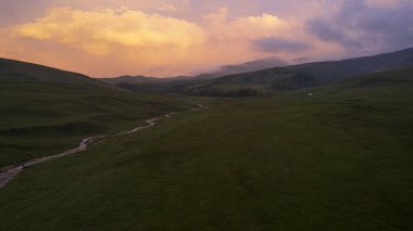 Yeşil tarlalı dağlarda pembe-mor gün batımı. Beyaz bir yurt var, bir dağ nehri akıyor. Kocaman bulutlar. Bir at sürüsü yürüyor. Çamur ve çadırın yanında bir SUV. Turistler toplanıyor