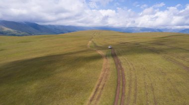 Beyaz bir SUV dağlarda tarlalardan geçiyor. Devasa beyaz bulutlardan, karlı dağ zirvelerinden ve yeşil tarlalardan oluşan bir dronun hava görüntüsü. Su birikintileri olan kirli bir off-road. Tekerleklerden damlıyor