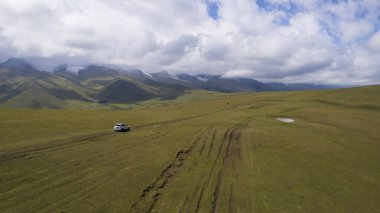 Beyaz bir SUV dağlarda tarlalardan geçiyor. Devasa beyaz bulutlardan, karlı dağ zirvelerinden ve yeşil tarlalardan oluşan bir dronun hava görüntüsü. Su birikintileri olan kirli bir off-road. Tekerleklerden damlıyor