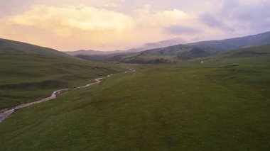 Yeşil tarlalı dağlarda pembe-mor gün batımı. Beyaz bir yurt var, bir dağ nehri akıyor. Kocaman bulutlar. Bir at sürüsü yürüyor. Çamur ve çadırın yanında bir SUV. Turistler toplanıyor