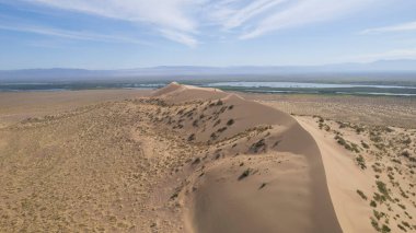 Bozkırın ortasında büyük bir kum tepeciği. Kocaman bir kum yığınının drone görüntüsü. Turistler yürüyor, manzaranın tadını çıkarıyorlar. Uzakta bir nehir akar ve çimenler büyür. Beyaz bulutlu gökyüzü
