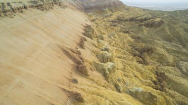 Renkli yüksek dağlar ve balçıktan yapılmış bir kanyon. Farklı taşları ve farklı renkleri olan büyük bir vadi. Kayaların duvarlarında kırmızı, turuncu, beyaz ve sarı çiçekler var. Bir turist yürüyor. Aktau