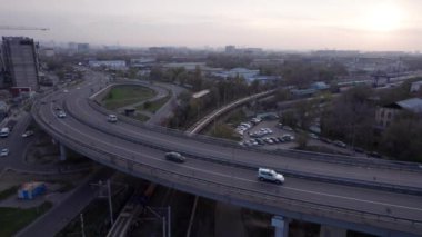 Şehirden geçen köprünün drone görüntüsü. Arabalardan kamyonlara farklı arabalar geçiyor. Serbest şerit. Gün batımında sisli bir şehrin arka planına karşı. Fabrikanın bacaları tütüyor.