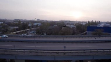 Şehirden geçen köprünün drone görüntüsü. Arabalardan kamyonlara farklı arabalar geçiyor. Serbest şerit. Gün batımında sisli bir şehrin arka planına karşı. Fabrikanın bacaları tütüyor.