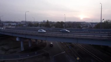 Şehirden geçen köprünün drone görüntüsü. Arabalardan kamyonlara farklı arabalar geçiyor. Serbest şerit. Gün batımında sisli bir şehrin arka planına karşı. Fabrikanın bacaları tütüyor.