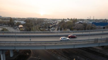 Şehirden geçen köprünün drone görüntüsü. Arabalardan kamyonlara farklı arabalar geçiyor. Serbest şerit. Gün batımında sisli bir şehrin arka planına karşı. Fabrikanın bacaları tütüyor.