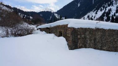An old stone prison or hut in the mountains. Snowy mountains, forest and clouds against a blue sky. Tall dry bushes peek out from under the snow. Birch and spruce trees are visible in the distance.