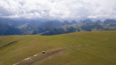 Beyaz bir SUV dağlarda tarlalardan geçiyor. Devasa beyaz bulutlardan, karlı dağ zirvelerinden ve yeşil tarlalardan oluşan bir dronun hava görüntüsü. Su birikintileri olan kirli bir off-road. Tekerleklerden damlıyor