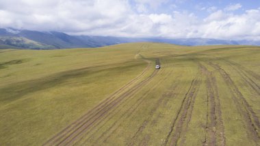 Beyaz bir SUV dağlarda tarlalardan geçiyor. Devasa beyaz bulutlardan, karlı dağ zirvelerinden ve yeşil tarlalardan oluşan bir dronun hava görüntüsü. Su birikintileri olan kirli bir off-road. Tekerleklerden damlıyor