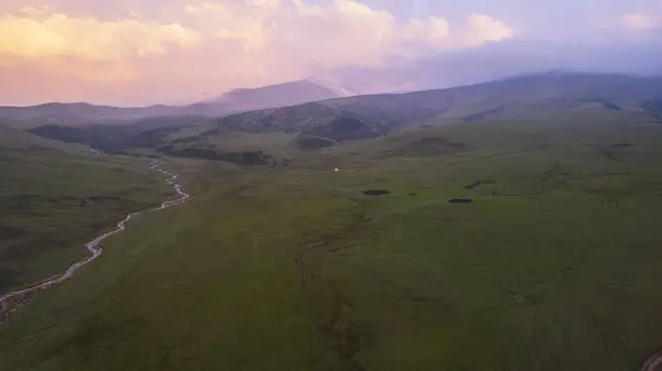 Pink-purple sunset in the mountains with green fields. There is a white yurt, a mountain river runs. Huge clouds. A herd of horses is walking. An SUV next to the mud and a tent. Tourists gather