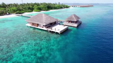 Turkuaz berrak su ve yeşil Maldivler adası. Okyanusun ve resifin üst manzarası. Palmiye ağaçları adada yetişir. Bungalovlar ve villalar var. Kumsalda beyaz kum. Balıklar görünür.