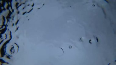 Suyun altından gökyüzüne ve yağmura bak. Damlalar suyun yüzeyine düşer ve su çemberleri oluşturur. Gri bulutlar, gökyüzünden gelen ışık ve damlalar görülebilir. Sıra dışı bir bakış.