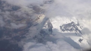 Beyaz bulutların ve karlı dağların düzlem penceresinden görünen manzara. Bir uçağın gümüş kanadı. Tepeler karla kaplı. Kahverengi kayalar. Bulutların gölgesi yere düşüyor.
