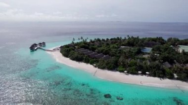 Turkuaz berrak su ve yeşil Maldivler adası. Okyanusun ve resifin üst manzarası. Palmiye ağaçları adada yetişir. Bungalovlar ve villalar var. Kumsalda beyaz kum. Balıklar görünür.