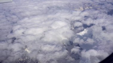 Beyaz bulutların ve karlı dağların düzlem penceresinden görünen manzara. Bir uçağın gümüş kanadı. Tepeler karla kaplı. Kahverengi kayalar. Bulutların gölgesi yere düşüyor.
