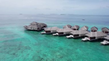 Okyanusun ortasında beyaz kumlu bir adanın insansız hava aracı görüntüsü. Turkuaz su, mercan resifi. Suda bungalovlar, sahilde villalar var. Her yerde palmiye ağaçları var. Maldivler