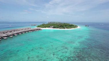 Okyanusun ortasında beyaz kumlu bir adanın insansız hava aracı görüntüsü. Turkuaz su, mercan resifi. Suda bungalovlar, sahilde villalar var. Her yerde palmiye ağaçları var. Maldivler
