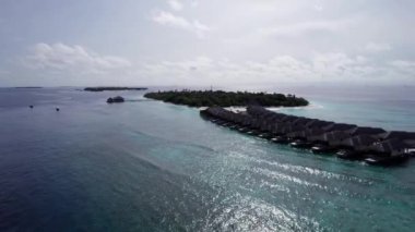 Sudaki bungalovun üst manzarası. Maldivler. Yeşil adanın yakınındaki lüks villalar. İnsansız hava aracının görüntüsü. Turkuaz su, güzel resif. Okyanusun yüzeyindeki güneş ışınları. Dalgalar