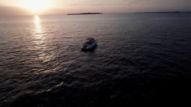 Deniz manzaralı turuncu gün batımı. İHA, yüksek hızlı beyaz bir teknenin etrafında uçuyor. Karanlık okyanus, suyun yüzeyindeki güneşin parlaklığı. Uzaktaki adalar görülebilir.