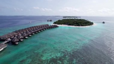 Sudaki bungalovun üst manzarası. Maldivler. Yeşil adanın yakınındaki lüks villalar. İnsansız hava aracının görüntüsü. Turkuaz su, güzel resif. Okyanusun yüzeyindeki güneş ışınları. Dalgalar