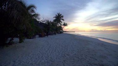 Beyaz kumlu tropik bir adada turuncu bir gün batımı. Yeşil palmiye ağaçları, ağaçlar ve çalılar. Gri bulutlar turuncu-kırmızıyla işaretlenmiştir. Sakin okyanus bir ayna gibidir. Bir köprü ve deniz kenarında bir ev