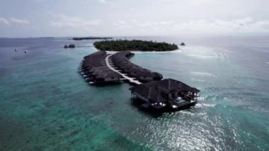 Sudaki bungalovun üst manzarası. Maldivler. Yeşil adanın yakınındaki lüks villalar. İnsansız hava aracının görüntüsü. Turkuaz su, güzel resif. Okyanusun yüzeyindeki güneş ışınları. Dalgalar