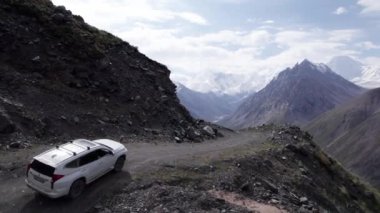 Dağlarda tehlikeli bir yol boyunca beyaz bir SUV sürüyor. Karlı tepelerin ve beyaz bulutların güzel bir manzarası var. Sağ taraftaki vadide bir uçurum var. Çimenler her yerde yetişir