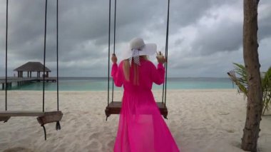 Pembe beyaz şapkalı bir kız sahilde salıncakta sallanıyor. Beyaz mercan kumu, bulutlu karanlık gökyüzü. Kız elinde bir kokteyl tutuyor. Turkuaz okyanus suyu ve yeşil palmiye ağaçları. Rahatlama