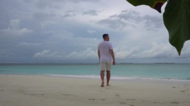 Beyazlı bir adam sahilde yürüyor ve heyecanlanıyor. Turkuaz berrak su, gökyüzünde kara bulutlar ve beyaz mercan kumu. Ağaçtan yapraklar. Kumda ayak izleri bırakıyor. Hafif meltem ve yağmur