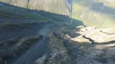 Vadideki dik uçurumlar boyunca bir FPV insansız hava aracına atlamak. Güneş ışınları tepenin arkasından geliyor. Büyük kayalar, yeşil çimenler ve vadiden aşağı akan bir nehir. Dağların arasında uçarak