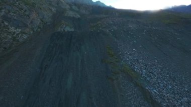 Vadideki dik uçurumlar boyunca bir FPV insansız hava aracına atlamak. Güneş ışınları tepenin arkasından geliyor. Büyük kayalar, yeşil çimenler ve vadiden aşağı akan bir nehir. Dağların arasında uçarak