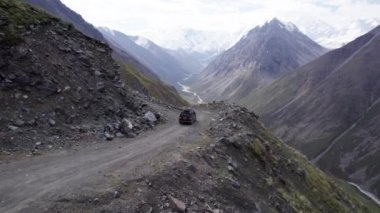 Siyah bir SUV dağlarda tehlikeli bir yol boyunca gidiyor. Karlı tepelerin ve beyaz bulutların güzel bir manzarası var. Sağ taraftaki vadide bir uçurum var. Çimenler her yerde yetişir