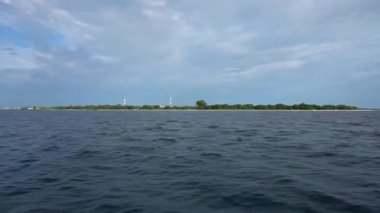 Yüzen bir tekneden okyanustaki bir adaya bak. Beyaz kum, yeşil ağaçlar. Baz istasyonları var. Alçak evler bazı yerlerde görülebilir. Gökyüzü bulutlarla kaplı. Okyanusu aşmak