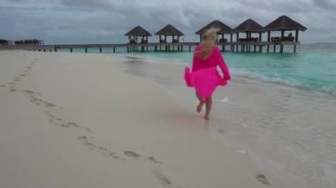 Pembe elbiseli bir kız kumsalda koşuyor. Kumaş rüzgarda dalgalanıyor, dalgalar hafifçe alıyor. Turkuaz okyanus suyu. Çardağın maliyeti. Temiz kum, dalgalar izleri temizler.