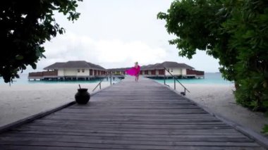 Pembe elbiseli bir kız bir adada köprüden geçiyor. Yeşil ağaçlar, sudaki villalara giden ahşap bir köprü. Turkuaz okyanus suyu. Pembe elbiseler rüzgarda dalgalanıyor. Bir İHA 'dan ateş etmek